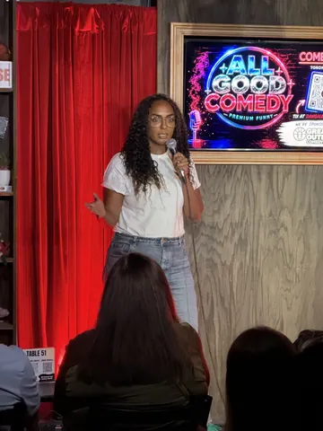 A comedian in a white top speaking beside the red stage curtains