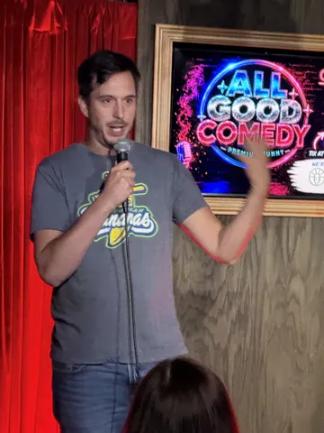 A comedian in a grey T-shirt gesturing with a microphone on stage