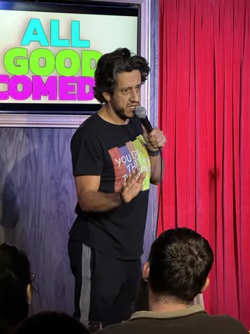 A comedian in a black T-shirt performing under the colourful All Good Comedy logo
