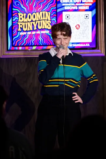 A comedian in a green and navy striped sweater performing beneath the Bloomin Funyuns screen