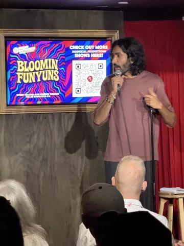 A comedian in a mauve T-shirt speaking into a microphone with the audience in the foreground