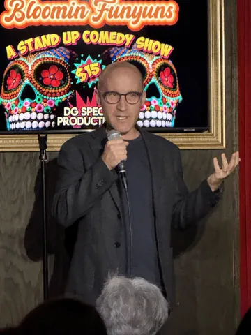 A comedian in a dark jacket gesturing beside a Bloomin Funyuns screen