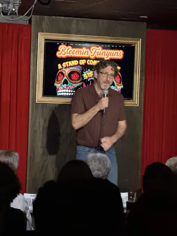 A comedian in a brown T-shirt performing beside red stage curtains