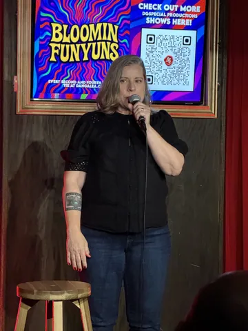 A comedian in a black shirt and jeans beside a stool on stage