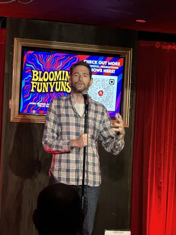 A comedian in a checked shirt performing under colourful lights