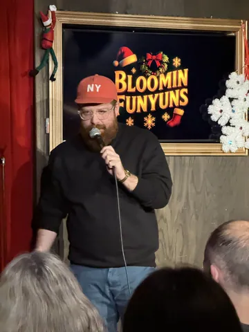 A comedian in a red cap holding a microphone in front of a Bloomin Funyuns screen