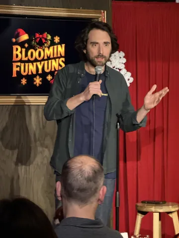A comedian in a dark jacket gesturing during a stand-up set