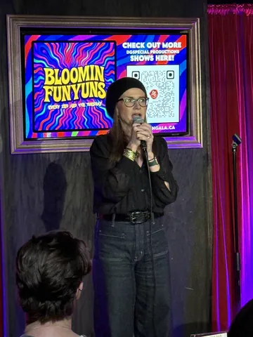A comedian wearing glasses and a black jacket holding a microphone