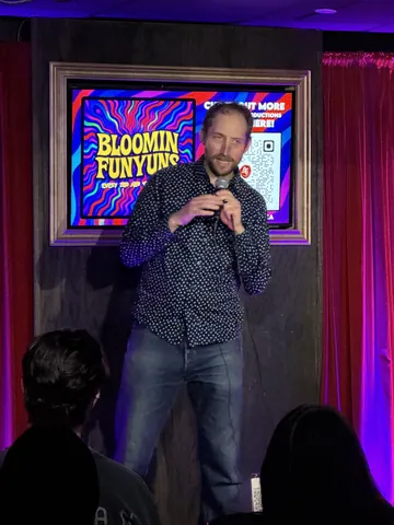 A comedian in a dark patterned shirt gesturing beside a stool