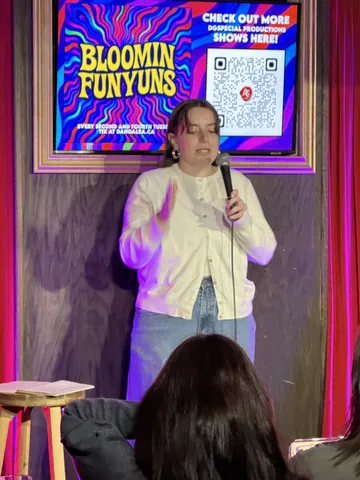 A comedian in a cream cardigan performing beneath the colourful show screen