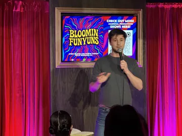 A comedian in a dark T-shirt holding a microphone beside the red curtains