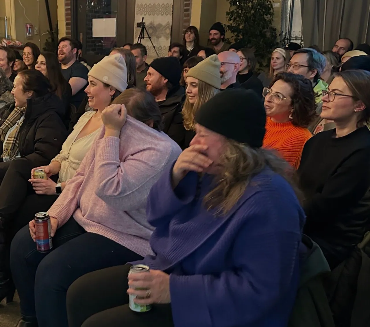 A packed indoor audience laughing, with a person pulling a lavender sweater over their face
