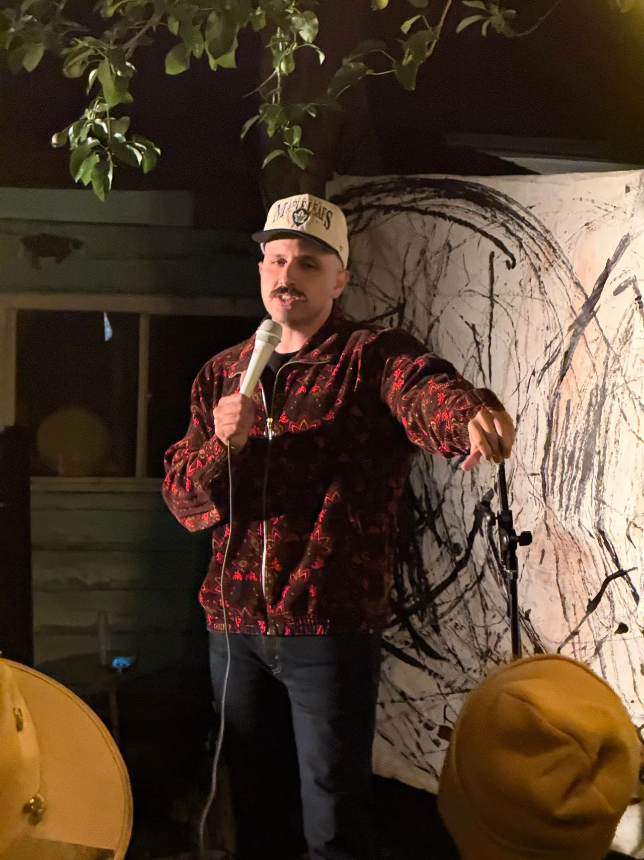 A performer in a red plaid jacket entertaining an outdoor audience beside a garden shed
