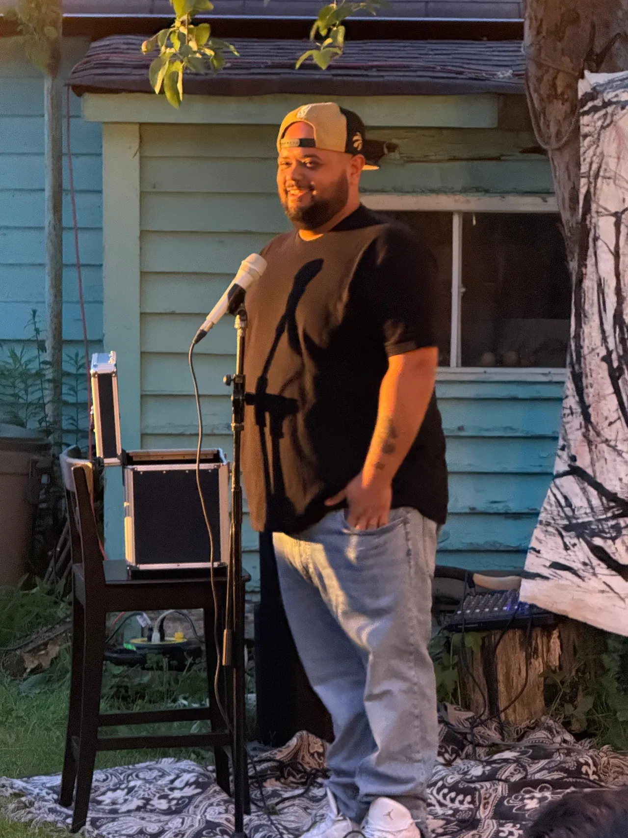 A comedian at a microphone beside a blue garden shed, with a stool and patterned backdrop