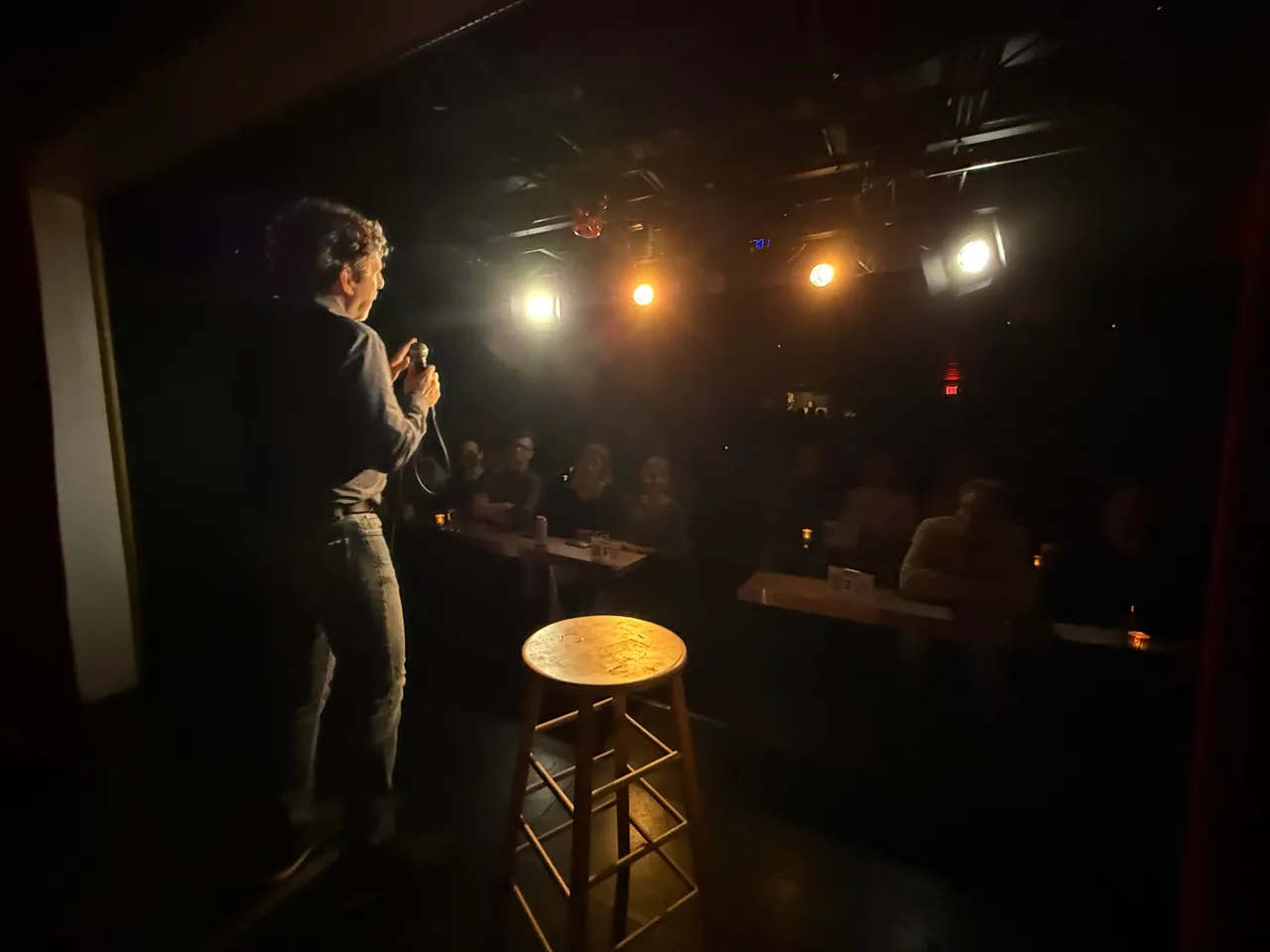A comedian seen from the side of the stage, facing a warmly lit audience with a wooden stool in the foreground