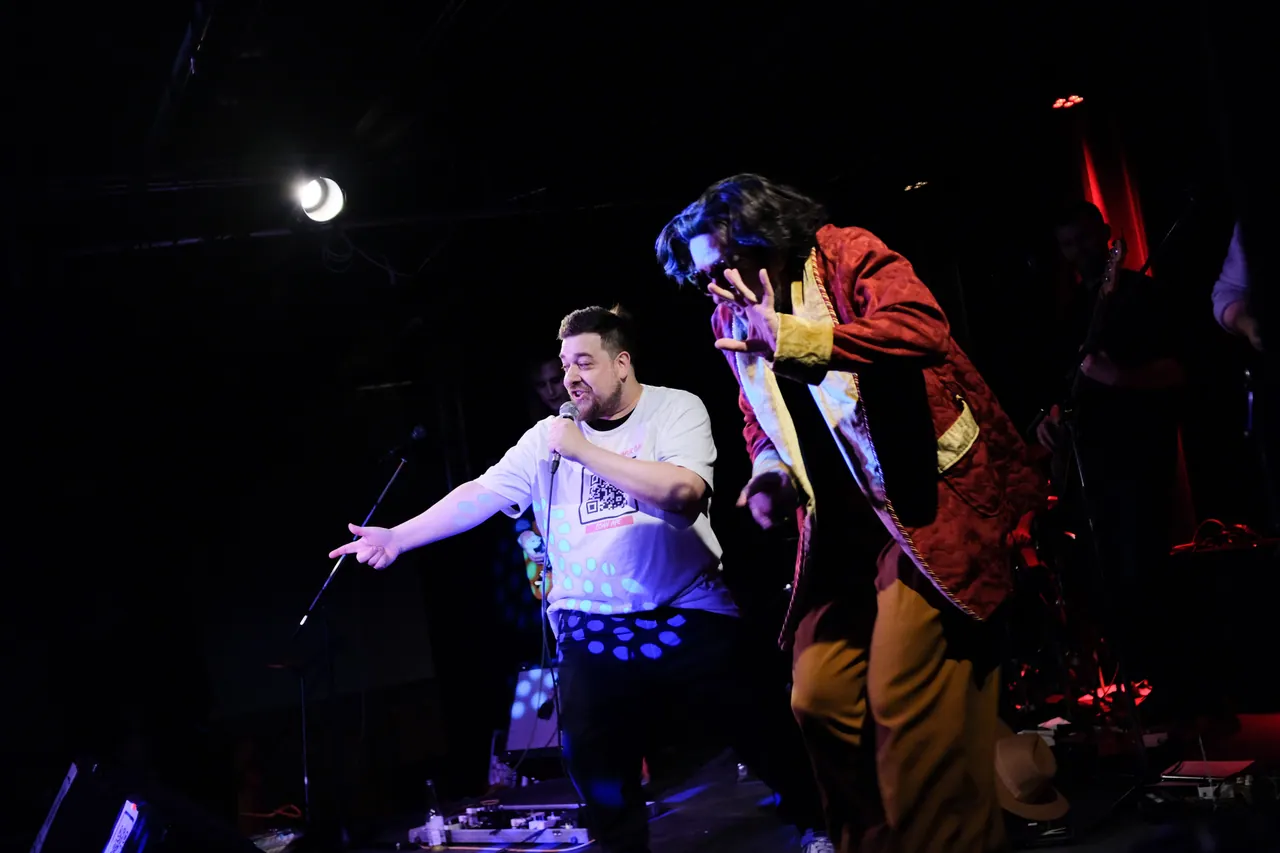 Two performers onstage, one wearing a red and gold theatrical coat
