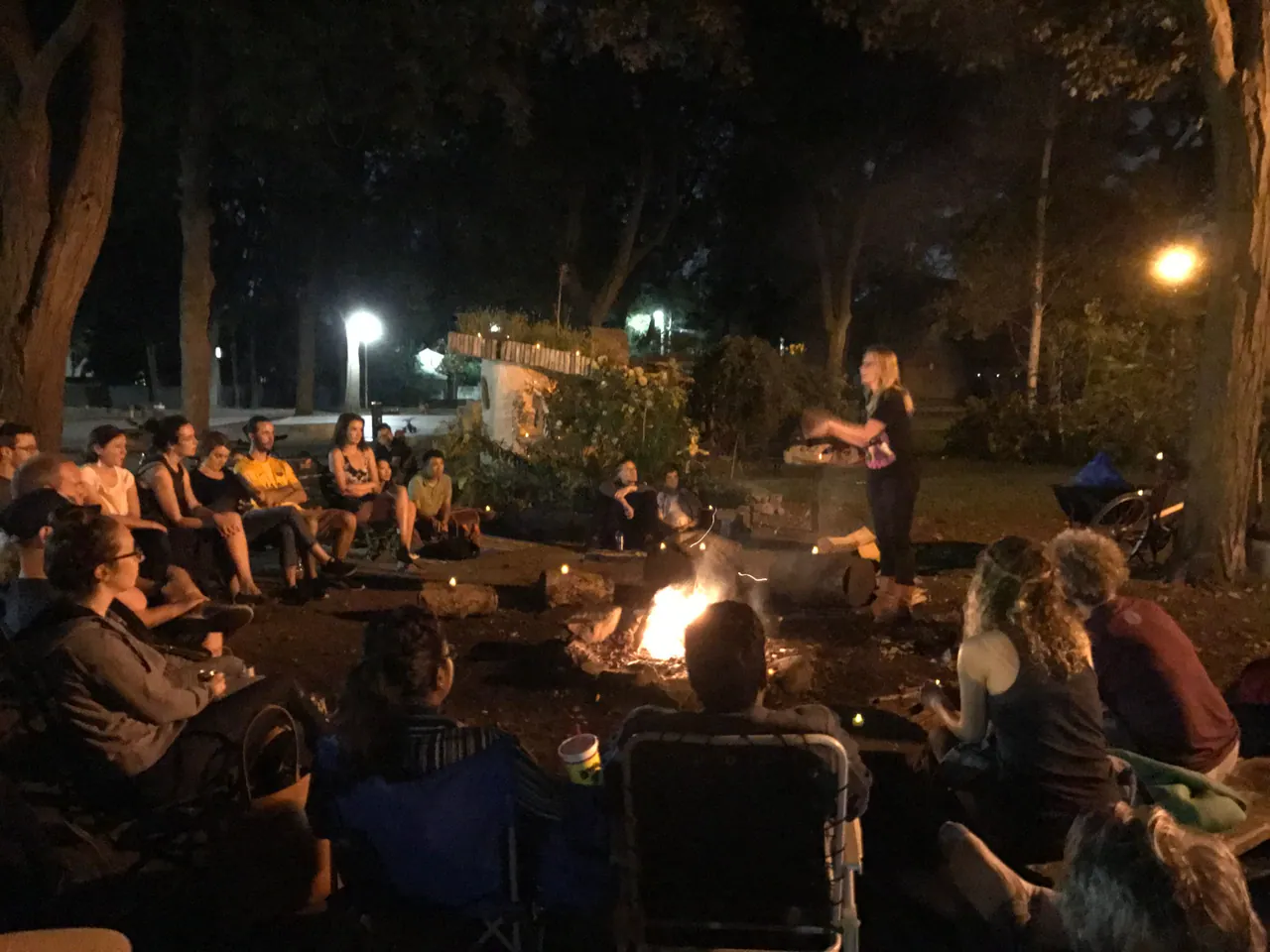 A performer entertaining an outdoor audience gathered around a fire beneath tall trees at night