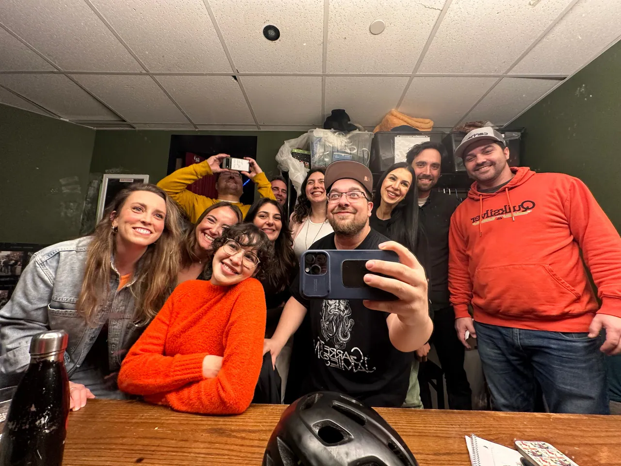 A cheerful backstage group posing for a mirror selfie, with bright orange sweaters in the foreground