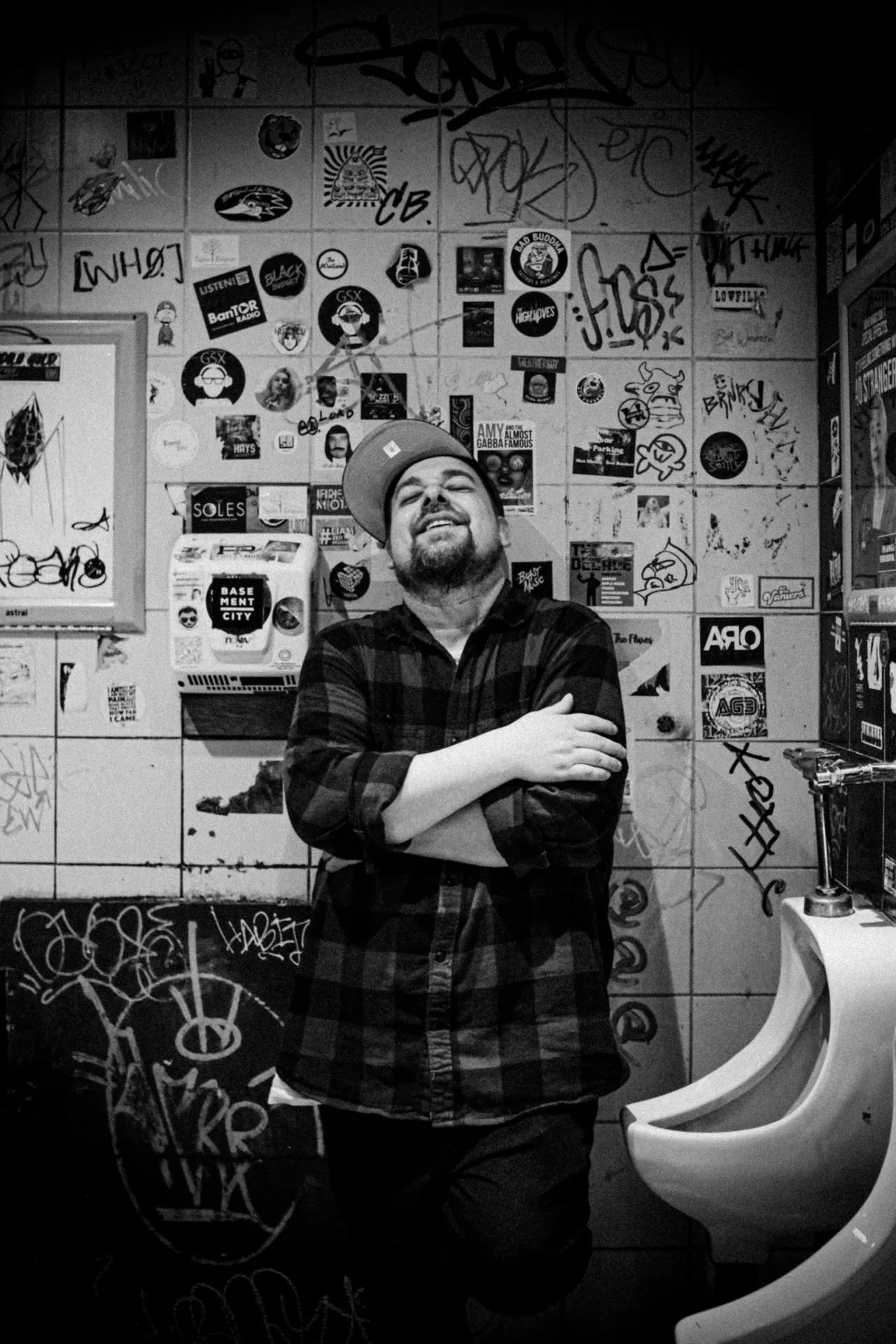 A black-and-white portrait of a smiling performer in a cap and plaid shirt against a wall of stickers