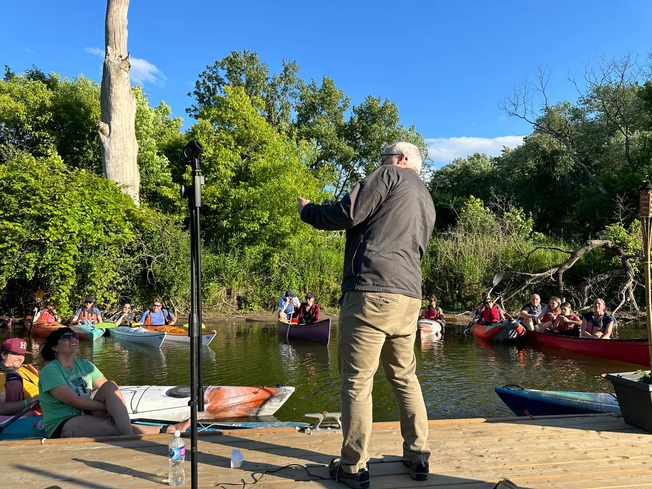 A performer on a wooden dock facing an audience in canoes and kayaks on the river