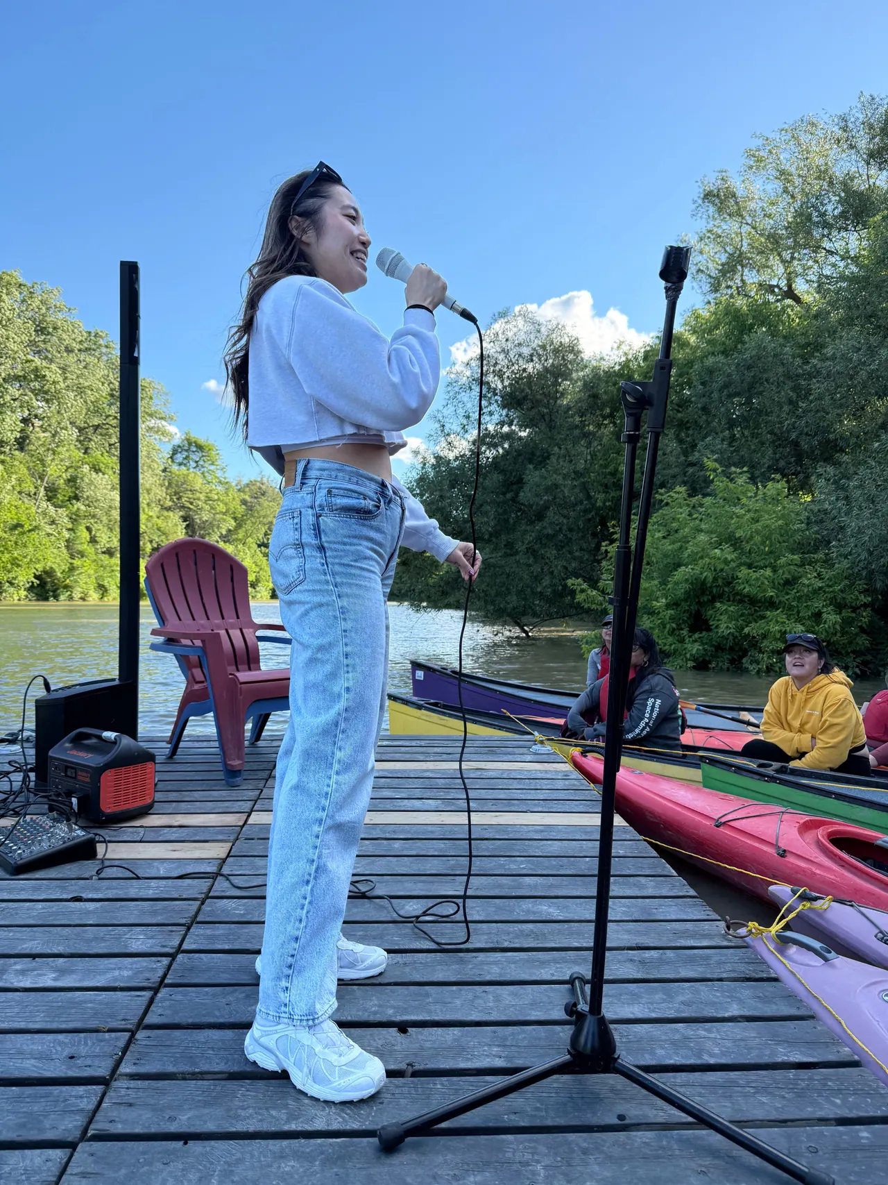 A comedian performing on a dock beside colourful kayaks on a sunny day