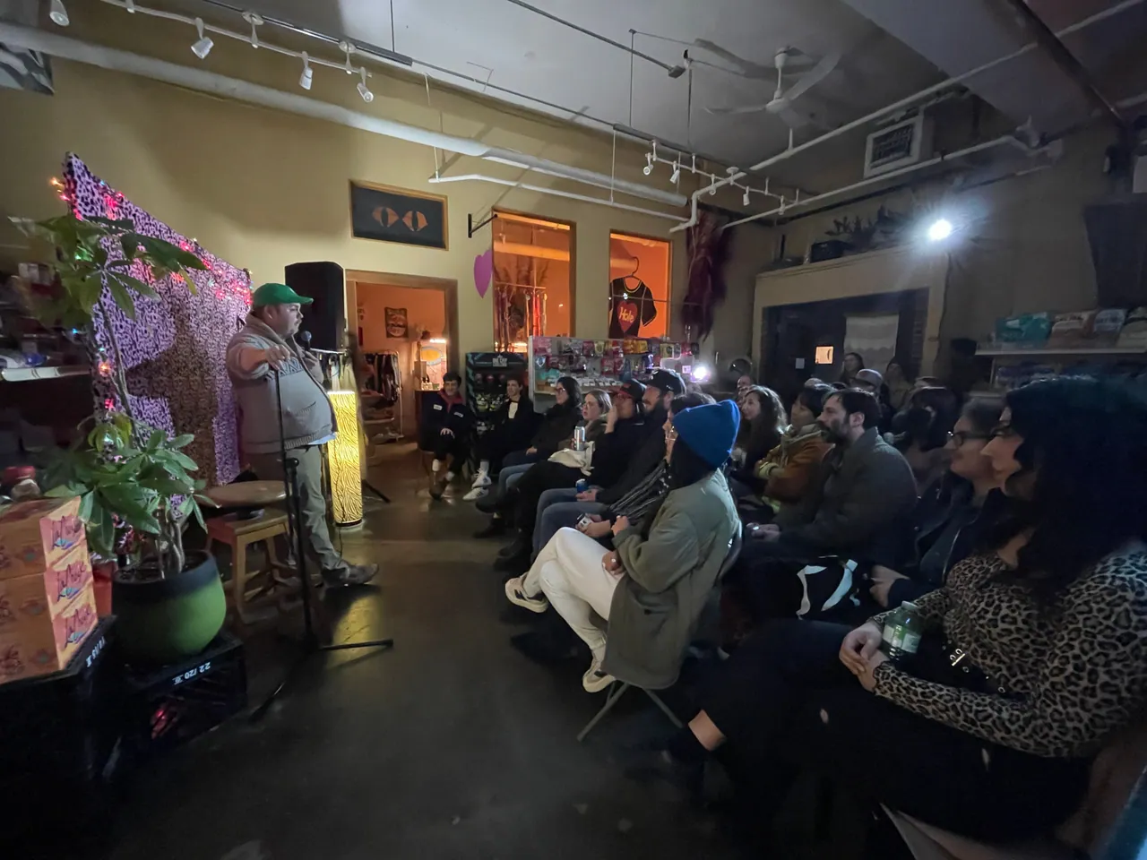 A comedian with a microphone performing for a seated audience inside a neighbourhood shop
