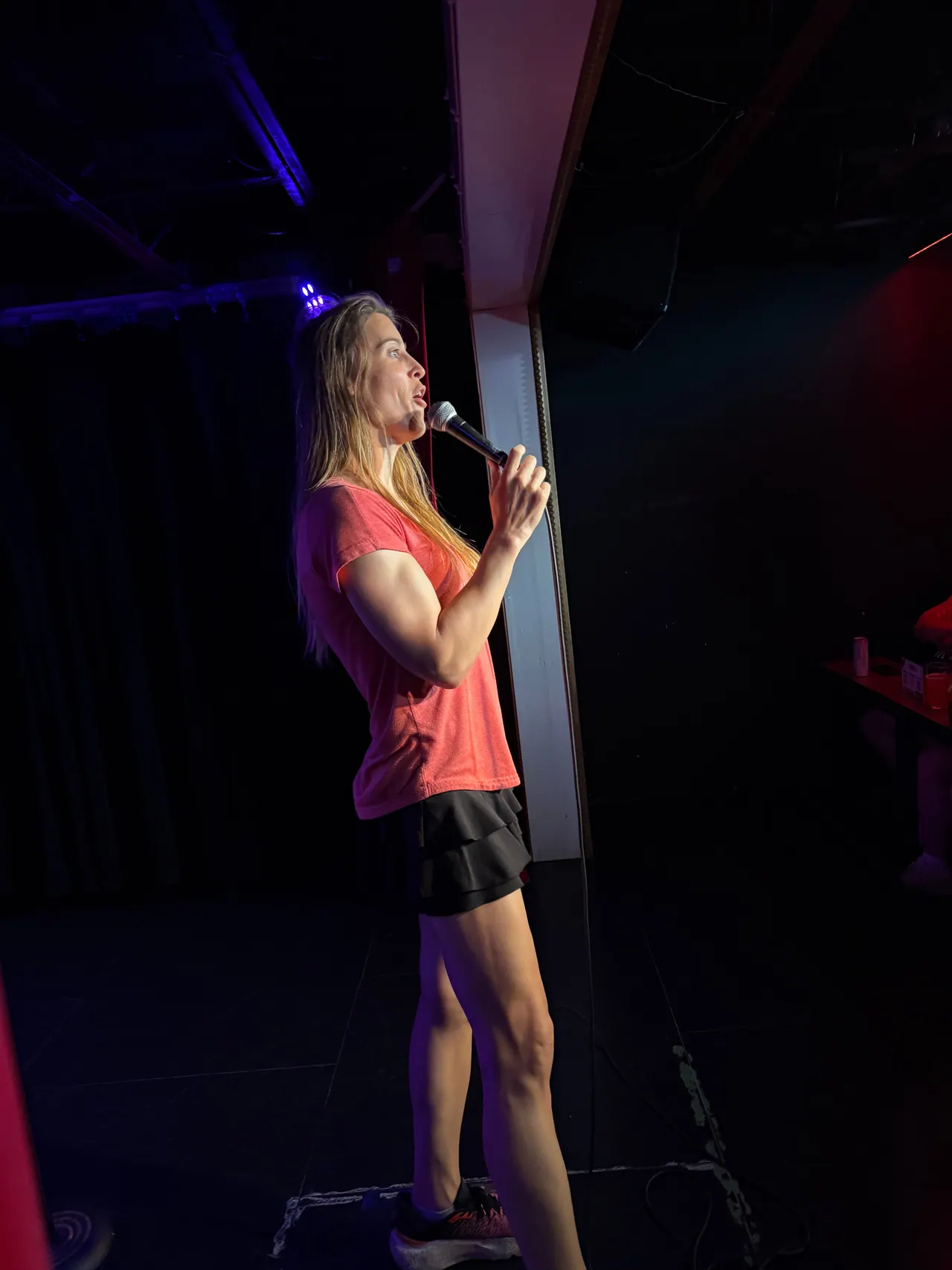A performer in a pink top and black skirt holding a microphone on a dark stage