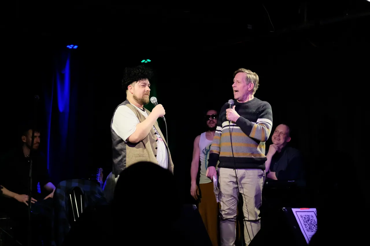 Two performers holding microphones with a band on the stage behind them
