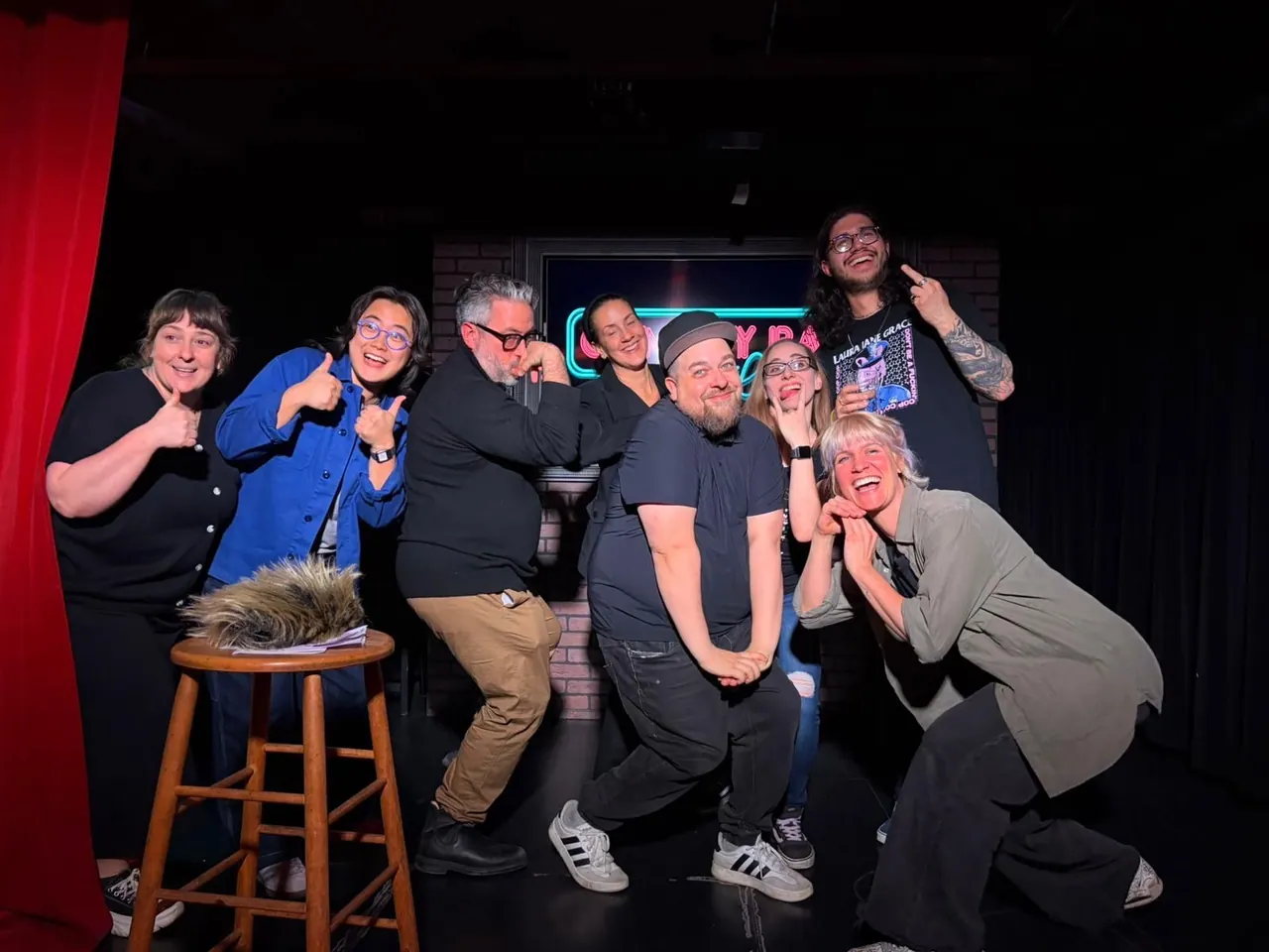 Seven performers making playful poses together on a comedy stage