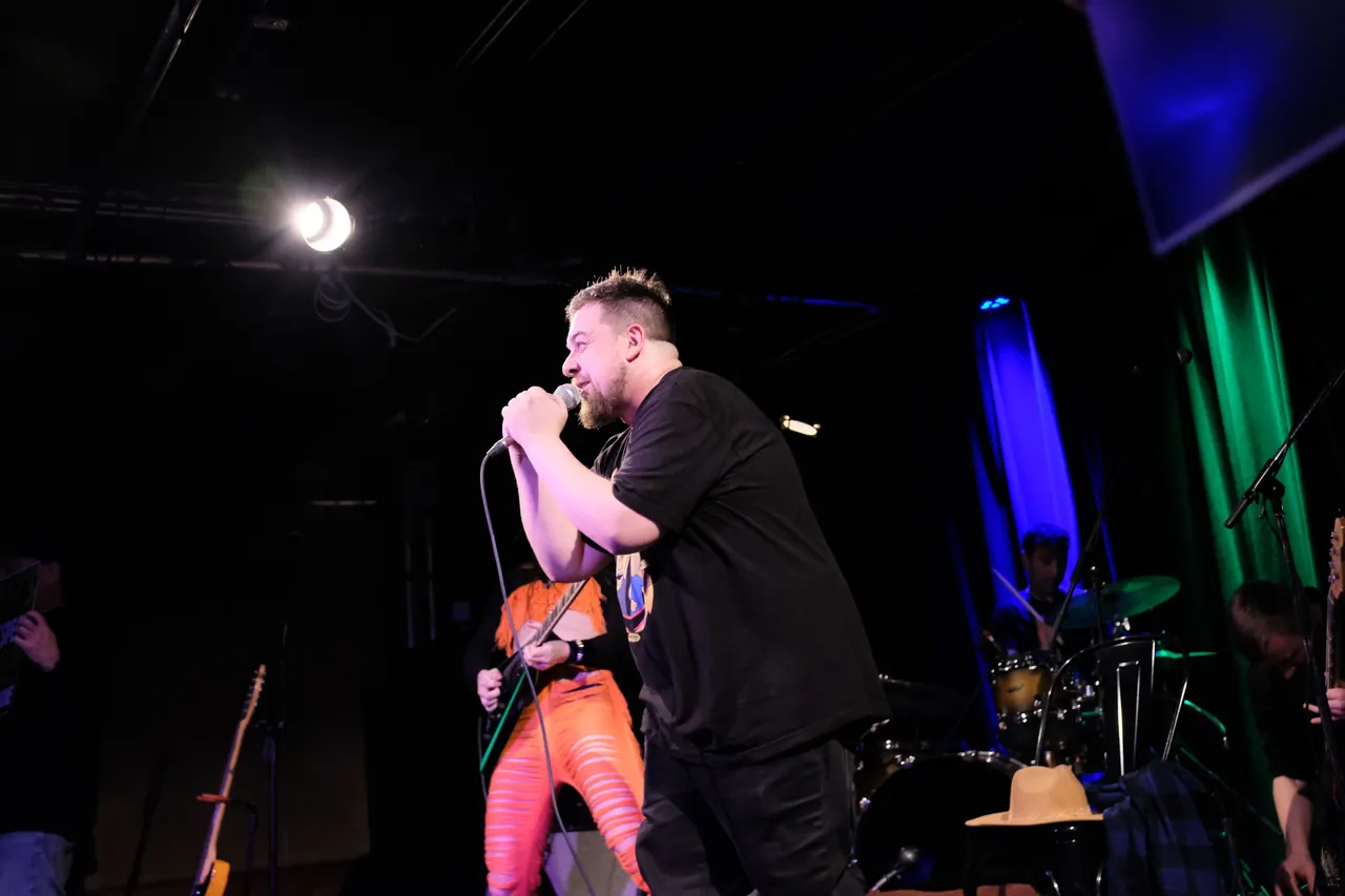 A performer at the microphone with a guitarist and drummer under coloured stage lights