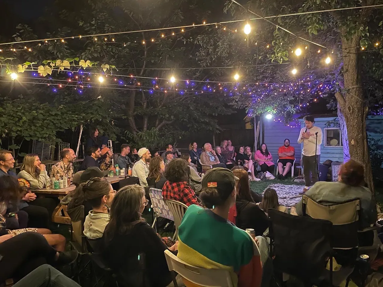 A backyard audience watching a comedian beside a blue shed beneath glowing string lights