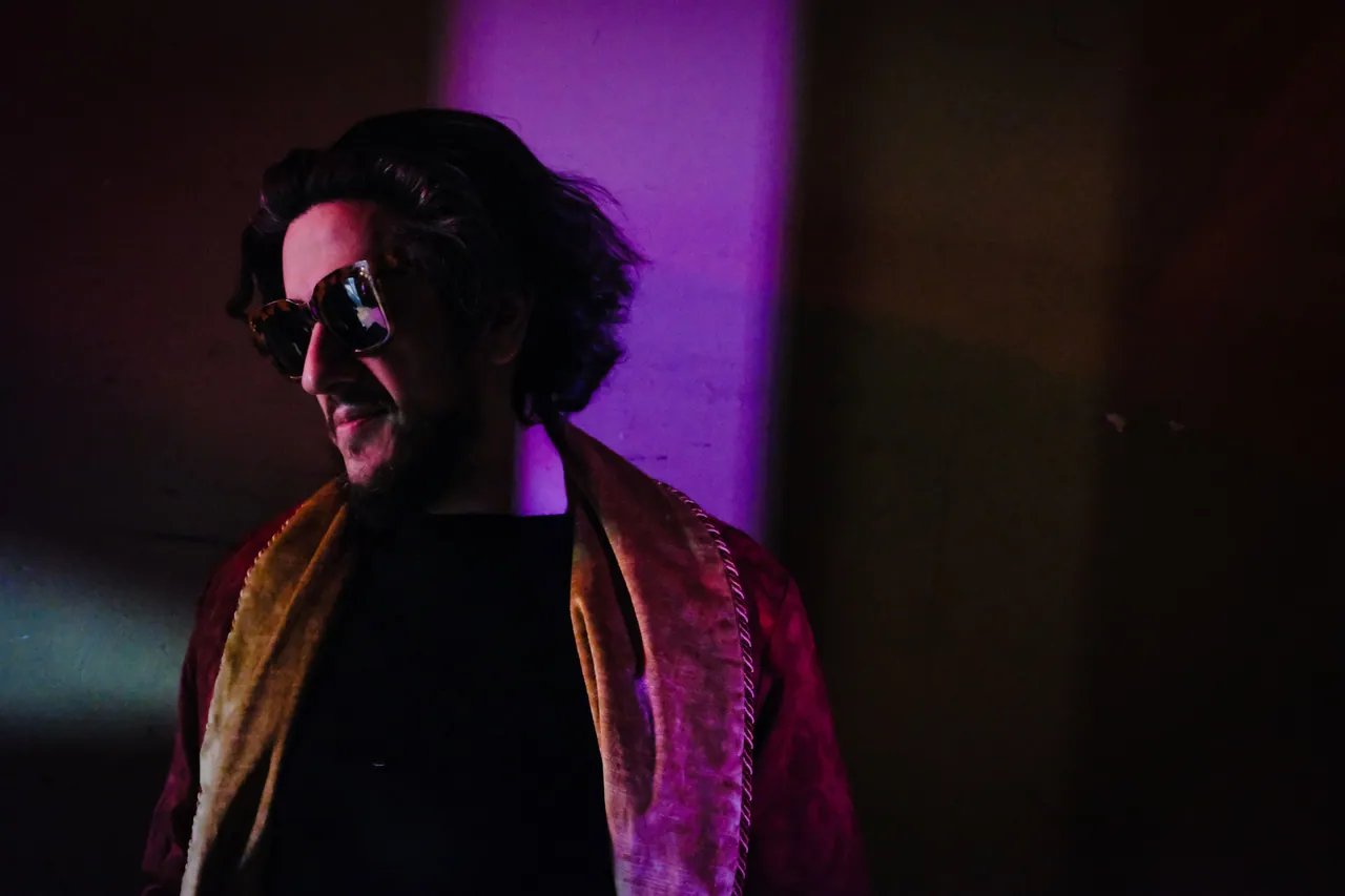 A performer in sunglasses and a red velvet coat standing under purple light